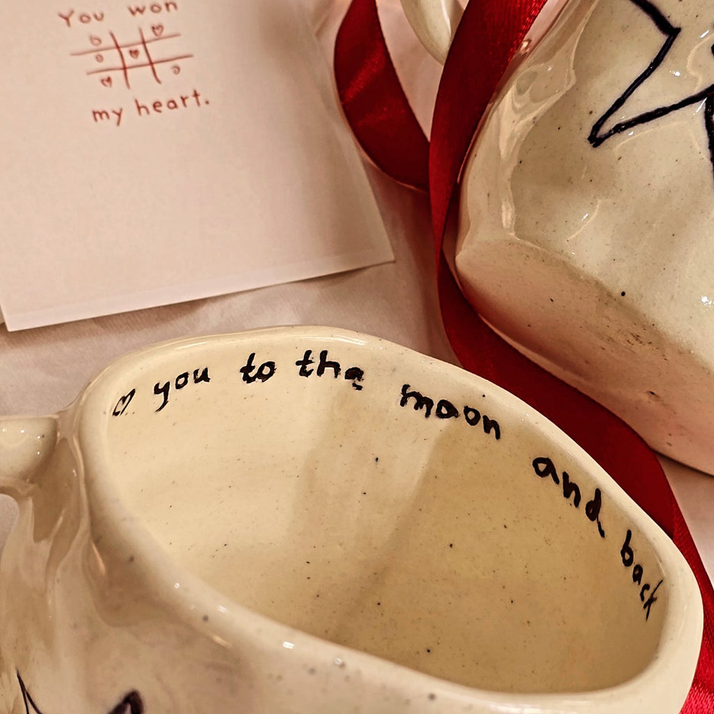 handmade valentines day ceramic mug by pastel jade ceramic studio with text love you to the moon and back and doodle of two stars holding hand in blue color  pastel jade in lahore pakistan all products are foodgrade and oven safe 