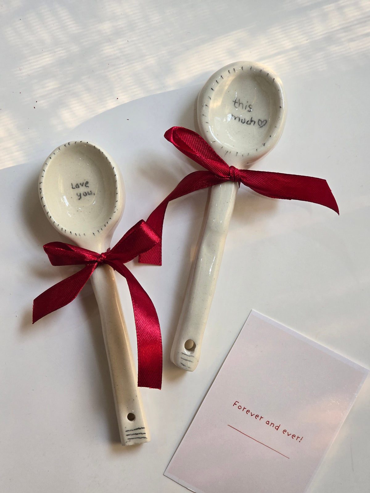 Two white ceramic spoons with red ribbon ties and text on the scoops, presented on a white surface with a card beside them.