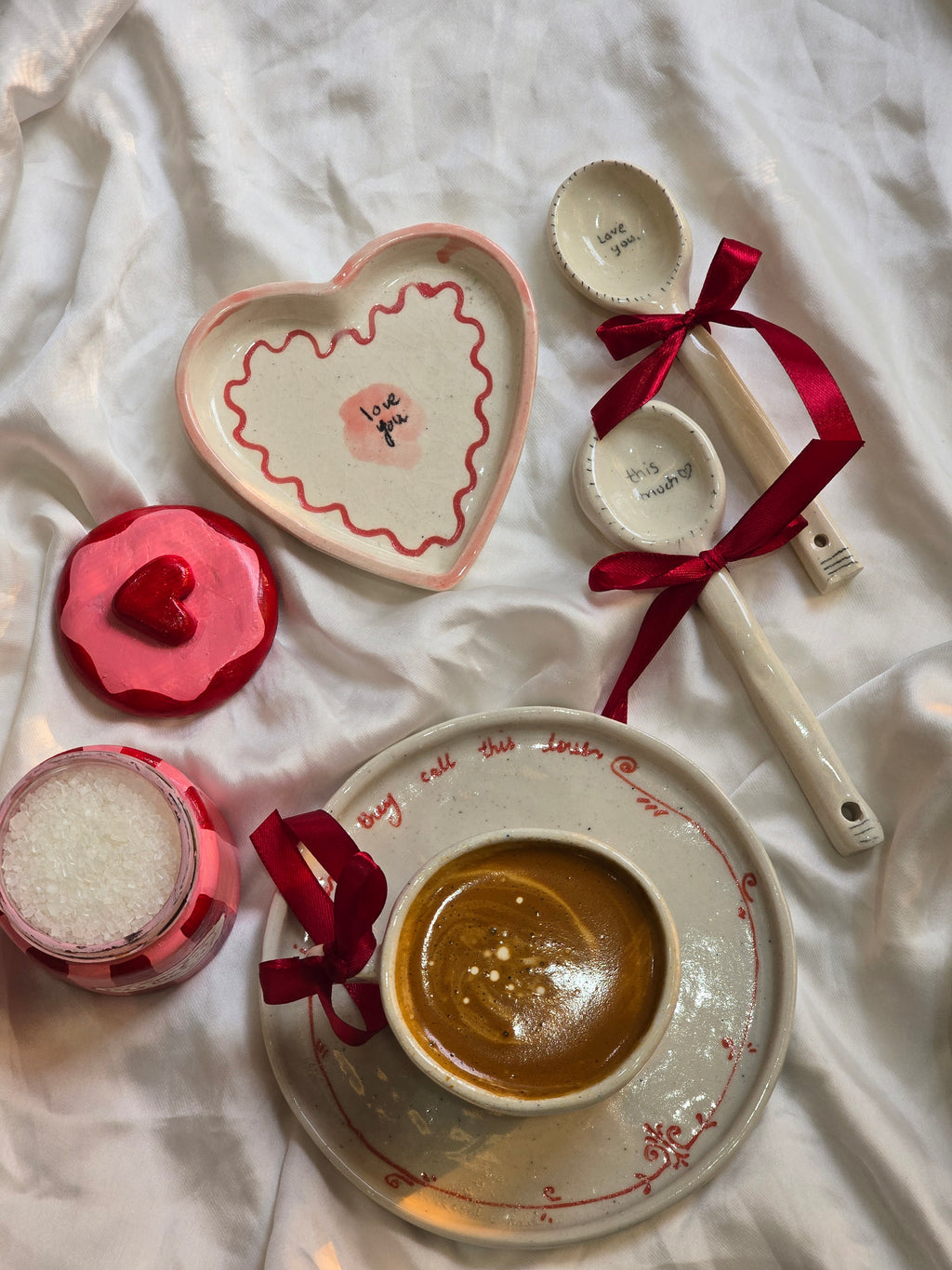 A collection of romantic-themed kitchenware including a heart-shaped bowl, a cup and saucer with heart patterns, a wooden spoon with a heart design, all arranged on a white surface.
