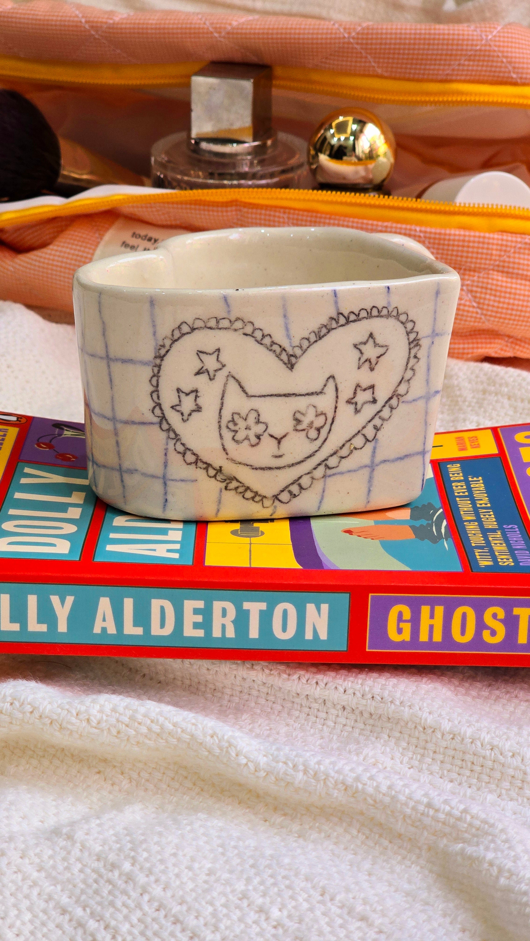 This image showcases a uniquely designed mug, featuring a whimsical depiction of a heart-shaped cat with whimsical elements. The mug is white, providing a neutral base that contrasts well with the colorful, hand-drawn style of the cat design.