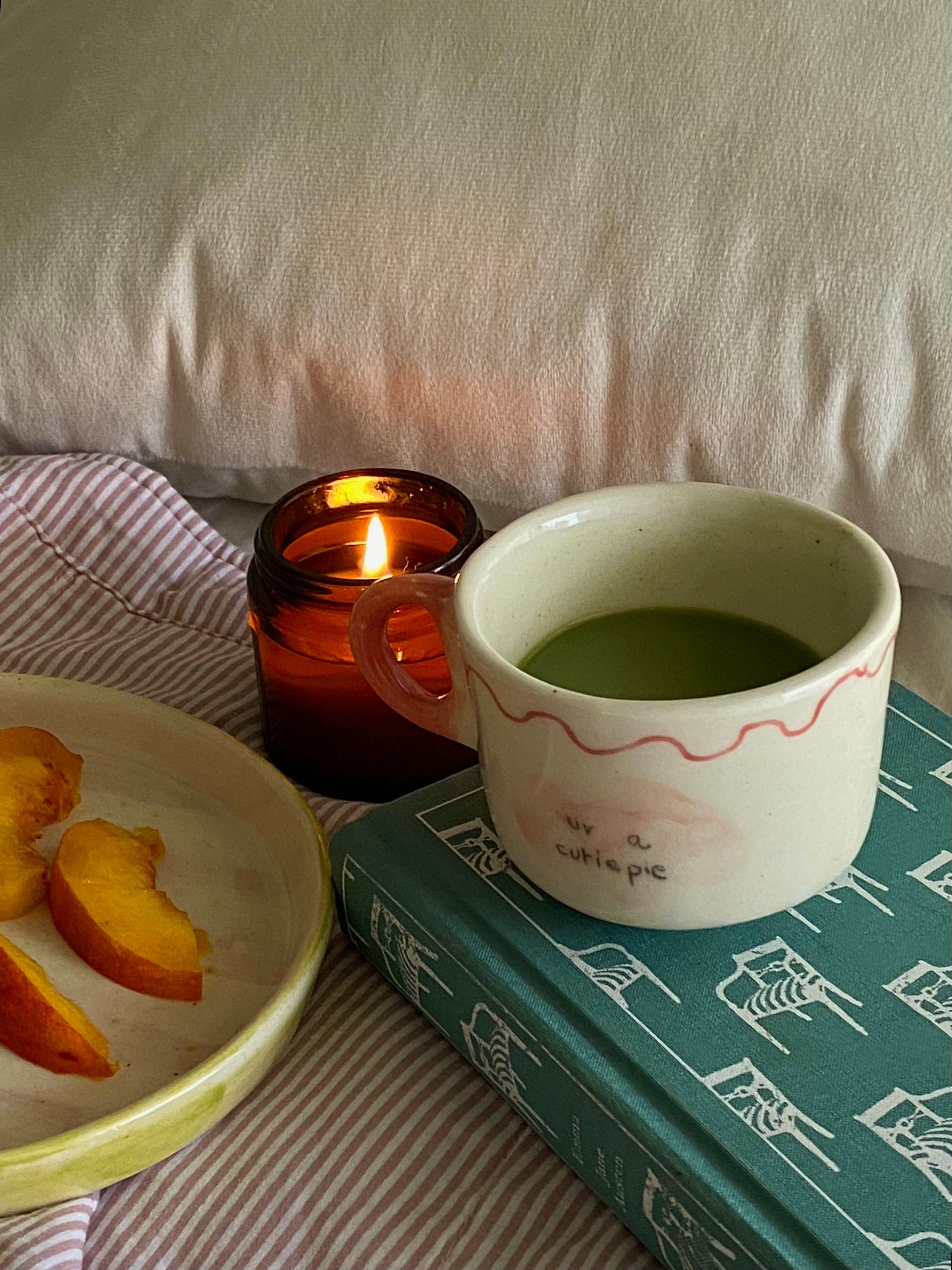 handmade ceramic macha cup wiuth love notes , gift perfect Cup of green tea macha on a wooden tray with a lit candle and peach slices, set against a striped fabric background.
