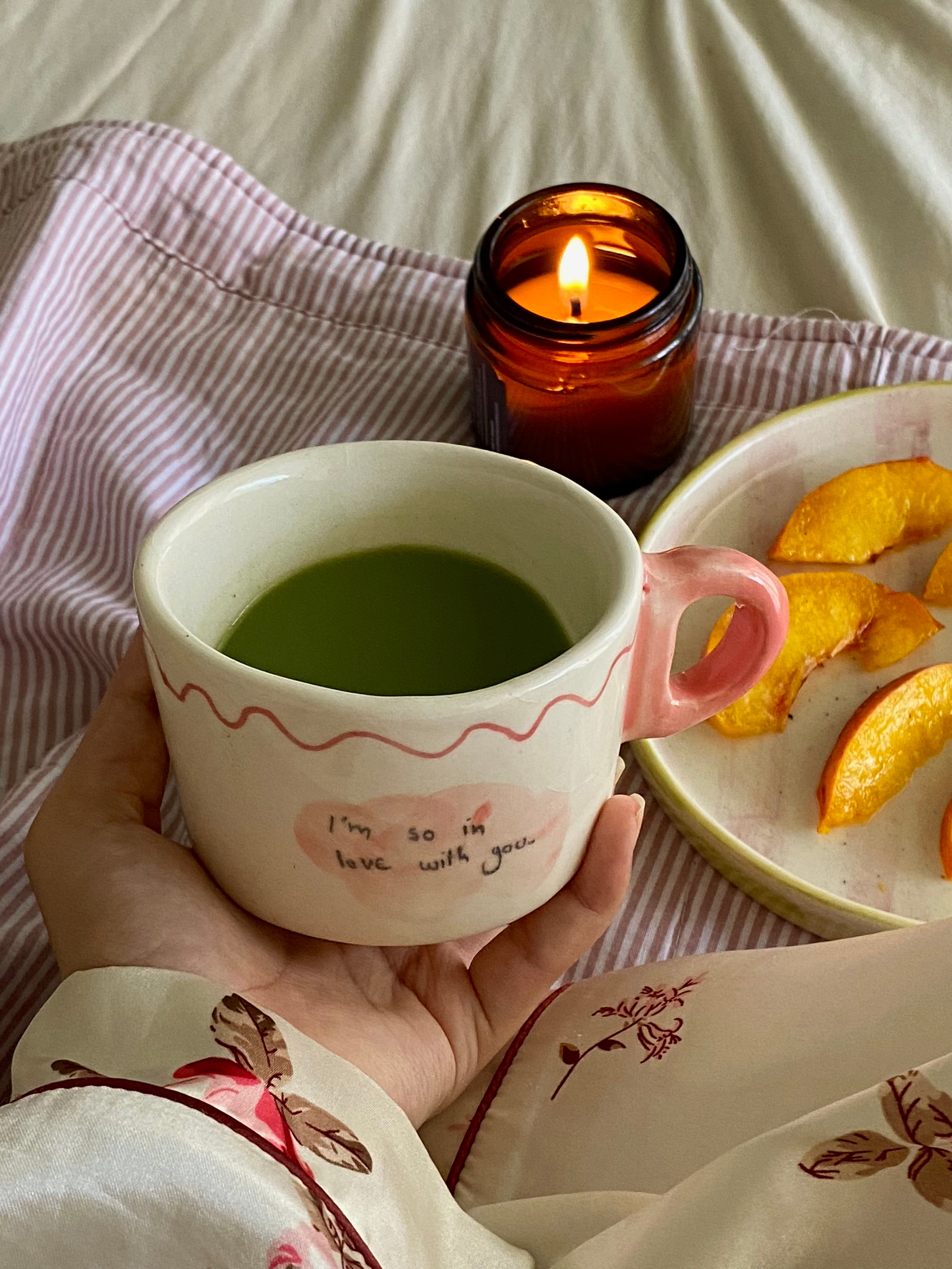 handmade ceramic macha cup wiuth love notes , gift perfect Cup of green tea macha on a wooden tray with a lit candle and peach slices, set against a striped fabric background.