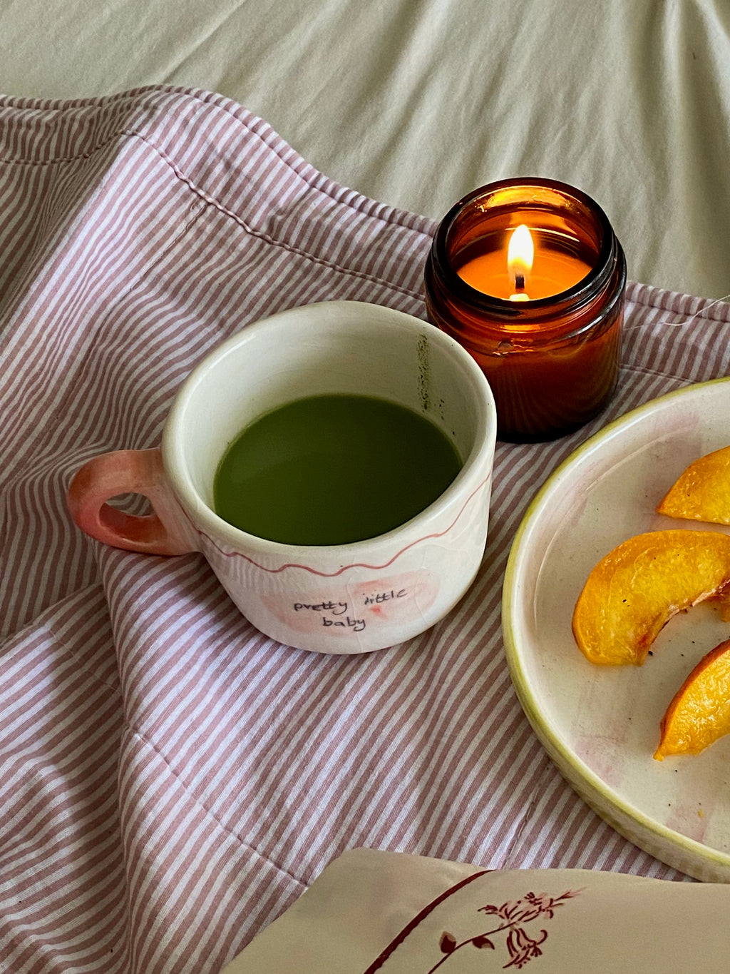 handmade ceramic macha cup wiuth love notes , gift perfect Cup of green tea macha on a wooden tray with a lit candle and peach slices, set against a striped fabric background.